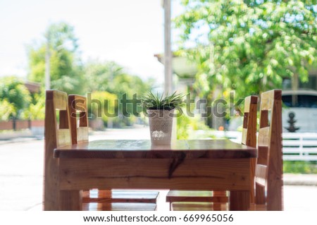 Brown wooden table with green flower in white pot in front of house with four chairs, soft focus.