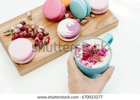 Woman hand hold blue cappuccino cup with roses petals. Beauty macarons on wood desk and white background