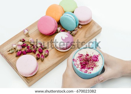 Beautiful french macarons on the desk. Woman hands holding a blue cup of cappuccino with roses petals