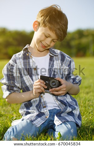 Vertical shot of a cheerful young boy relaxing outdoors in the park sitting on the grass holding vintage instant camera people photographer hobby retro work travelling children childhood happiness