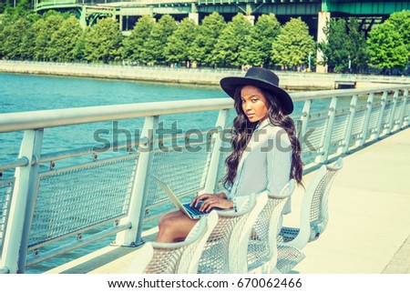 Young American woman with long hair traveling, working in New York in summer, wearing black sun hat, sitting on char at Riverside Park under sun, typing on laptop computer, looking up, thinking.
