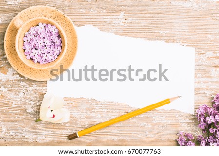 Piece of torn paper, pencil, beautiful cup with lilac flowers on old board. Place for your text.