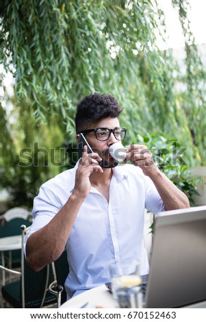Young attractive afro American businessman with glasses and laptop sitting in cafe bar and talking on cell phone. 