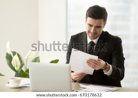 Smiling successful businessman holding document sitting at desk, reading good news in letter, reviewing beneficial lucrative contract, getting notification about bank loan or business credit approval  Royalty-Free Stock Photo #670178632