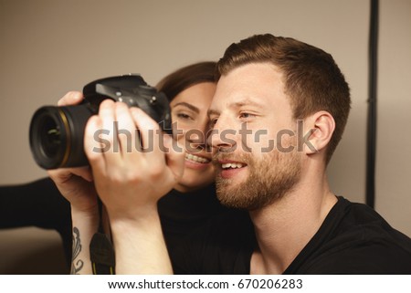 Indoor portrait of handsome bearded young male photographer holding professional camera, showing pictures from photo shooting to his attractive girlfriend at home. Selective focus on man's face.