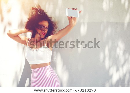 Beautiful young woman dressed in summer clothing photographing herself on smartphone camera. Attractive female making selfies on cellphone posing outdoors. Copy space area for advertise or information