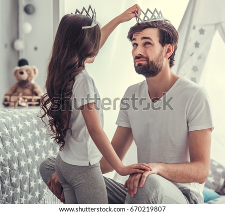 Cute little girl is putting a crown on her handsome young dad's head and smiling while they are playing together in child's room