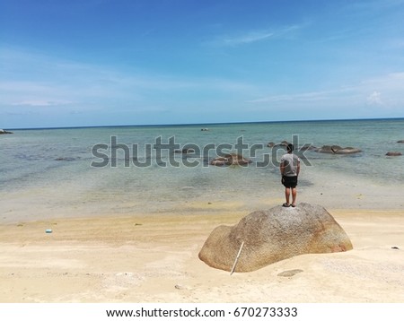 A picture of teenager standing on the big rock. looking at the clear water ocean seeing the diffrrent colour of seawater light and darker emerald green until thr end of touching blue sky  