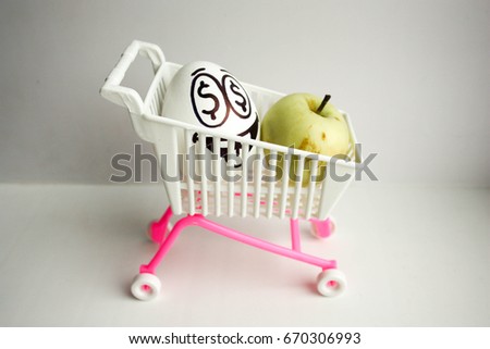 Concept of sales and income in dollars. A shopping basket with an apple and an egg with a painted face, eyes are dollars. To ride on wheels. Photo for your design.