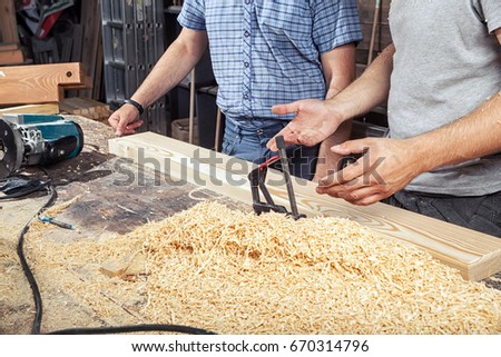Close-up as two men in a T-shirt and a checkered shirt stand near a wooden table and discuss how to process a wooden board with a milling machine, around a lot of wooden sawdust