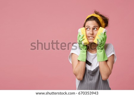 Funny charming young Caucasian housewife wearing yellow headband, green rubber gloves and striped apron, holding sponges at her face, having dreamy thoughtul look and smiling, posing at studio wall