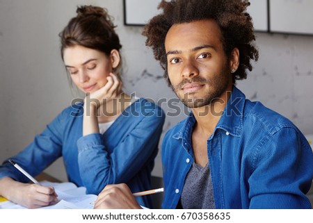 Two students working on common research project sitting at desk in library, making notes in copybook. Confident dark-skinned A-student helping cute shy girl to solve difficult mathemtical problem