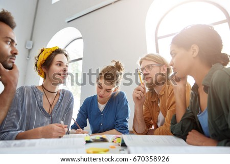 Indoor shot of group of multiracial students having meeting working together using books trying to write article about their studying at university sharing ideas with each other finding suitable words Royalty-Free Stock Photo #670358926
