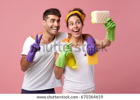 Funny couple having fun during doing housework and cleaning: smiling man hugging his wife who is making selfie with sponge. Cheerful male and female using sponge like smartphone making selfie together Royalty-Free Stock Photo #670364659
