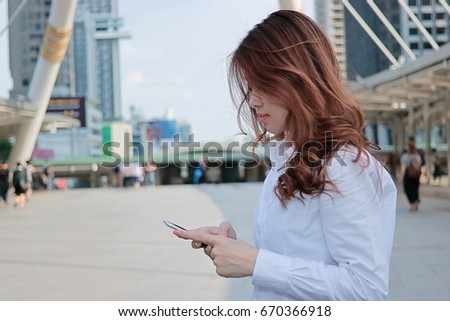 Side view of attractive young Asian business woman looking mobile smart phone in her hands at urban building city background.