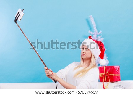 Enjoying christmas gifts. Woman in santa hat taking picture of herself using selfie stick. Indoor shot on blue background