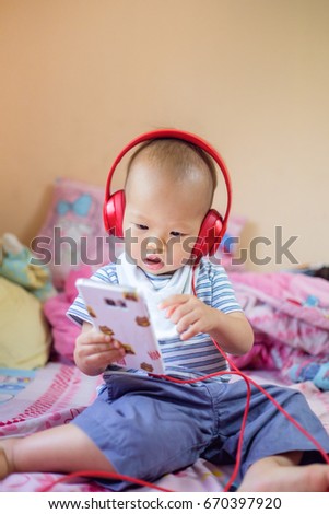 Cute little Asian 18 months / 1 year old baby boy child listening music with headphones from smartphone, Kid stand & Play on bed in bedroom at home, leisure & technology & internet addiction concept