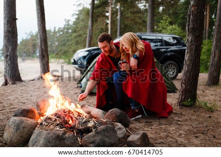 Beautiful couple, sitting, camping around the campfire. On the background with a green tent, camping mug with warm soup. Beside their off-road machine. The man flicks the fire with a stick.