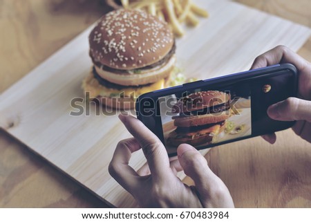 Treandy woman using her smartphone taking the fresh tasty big burger lunch photograph for sharing on her social community.Daily routine before eating every meal.Burgers are an American icon.