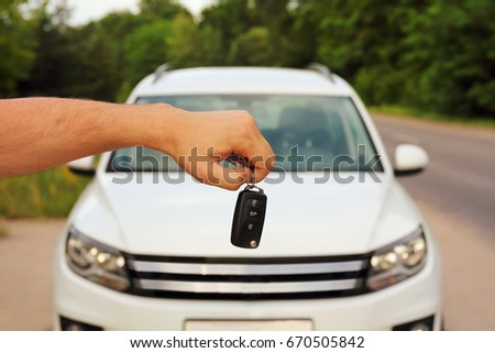 Male hand holding car keys on blurred background with new white car, green tree and road in summer outdoors.