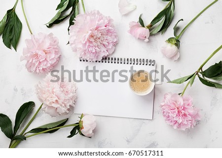Morning coffee cup for breakfast, empty notebook and pink peony flowers on white stone table top view in flat lay style. Woman working desk.