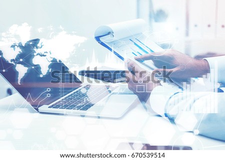 Close up of hands of a young businessman holding a clipboard and sitting near a laptop. Graphs and a world map. Toned image double exposure. Elements of this image furnished by NASA