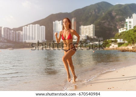Fit female athlete wearing bikini running on beach with sun shining in camera and hotel resort hills in background