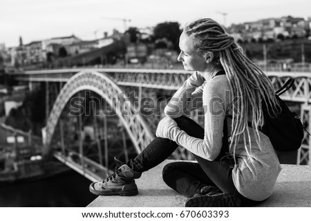 Black and white photo of young woman with dreadlocks sitting on the background of the Dom Luis I bridge in Porto, Portugal.