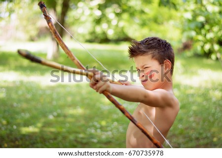 Adorable young boy playing native Indian with toy bow and arrow is aiming and about to shoot Royalty-Free Stock Photo #670735978