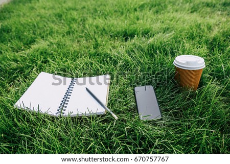 Close-up view of notebook with pencil, smartphone with blank screen and disposable coffee cup on green lawn