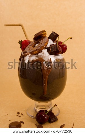 Chocolate indulgent extreme milkshake with brownie cake, strawberries, cherries, and a plastic straw with milk foam on top in a soft brown background