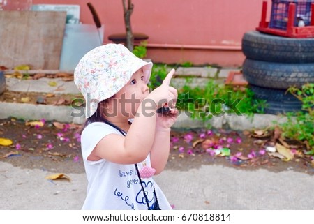 Portrait of adorable Asian girl wearing a hat looking up and pointing her right hand at a bird in the sky. The left hand holding the camera is taking a photo.