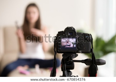 Close up photo of digital camera on tripod with young woman using cosmetics image on LCD back screen and blurred scene on background. Female video blogger records vlog, beauty vloger streaming online 
