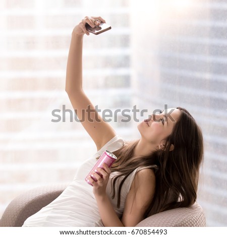 Young beautiful woman making selfie photo using cellphone while sitting in chair with can of refreshing beverage in hand. Pretty lady shooting herself for blog or social network post during vacation 