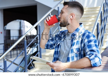 Close-up shot of bearded man with headphones listening to music on digital tablet, drinking coffee 