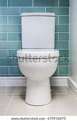 Clean new white toilet fitted against a tiled wall shot straight on. Royalty-Free Stock Photo #670936870