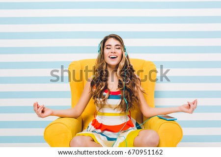 Joyful long-haired girl meditating while sitting in a lotus pose on blue striped background. Pretty young woman in colorful dress chilling in yellow armchair and listening relaxing music.