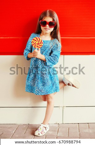 Portrait little girl child with a lollipop stick in leopard dress with sunglasses on a red background