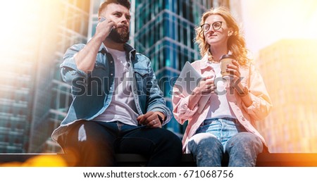 Two young business people are sitting outdoors. Hipster guy is talking on cell phone, girl is holding cup of coffee and tablet computer. In background, skyscraper in soft focus. Film effect. Lifestyle