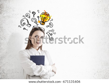 Portrait of an alert young woman with fair hair holding a blue book and standing with crossed arms near a concrete wall with question marks and bulb on it. Mock up