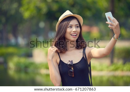 Pretty Vietnamese woman wearing straw hat standing at picturesque lake and taking selfie on smartphone, blurred background