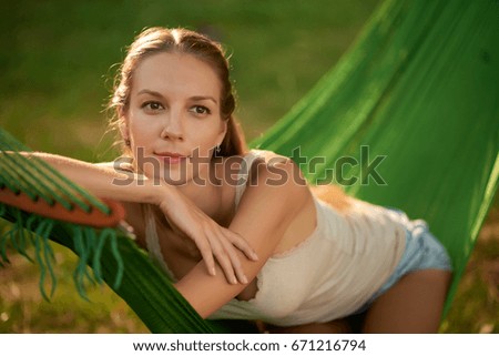 Portrait of beautiful young tourist looking away pensively while sitting in hammock, green lawn on background