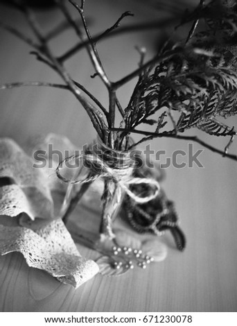 Dry tree branches in a transparent jar with a rope tied in a bow on it standing on wooden surface. Piece of lace fabric is lying next to it. Black and white picture