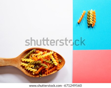 It's time for pasta dish, top view picture of multicolored uncooked fusilli consist of yellow, orange and green on wooden spoon above white, blue and pink  background.
