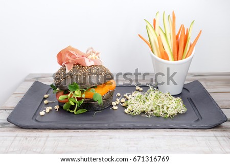 Healthy burger with hamon, tomatoes, micro greens and black wholegrain buns, vegetable sticks on black slate board over wooden background. Clean eating, dieting, detox food concept.