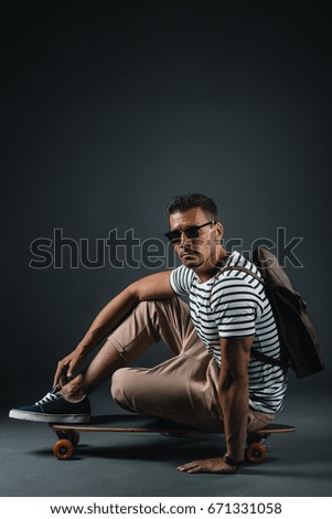 Handsome stylish man in sunglasses sitting on skateboard and looking at camera