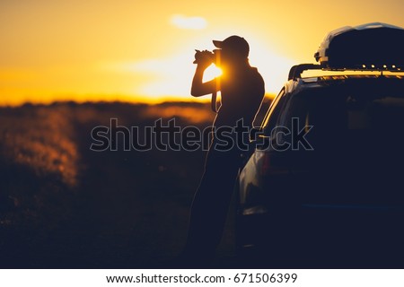 Nature Wildlife Photographer Working in the Field. Senior Photographer Taking Pictures on the Country Road While Supporting His Back on His Vehicle. Scenic Countryside Sunset.