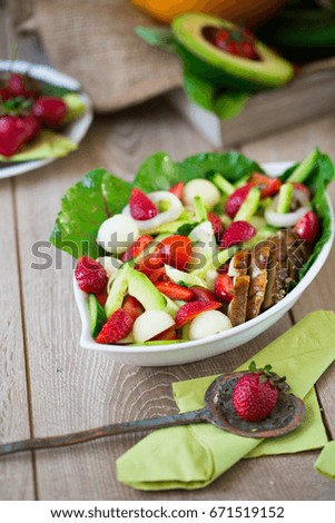 Oval white plate with warm chicken salad with strawberries, lemon, leek, tomatoes, peas, mangold leaves, avocado, melon balls, cucumber slices and white cheese on wooden table with fork and spoon