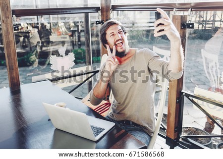 Happy attractive guy making selfie showing victory gesture and smiling. Outdoor cafe background