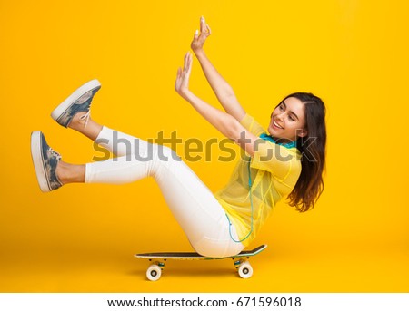Young woman sitting on skate with hands and legs up riding in studio on yellow background.  Royalty-Free Stock Photo #671596018